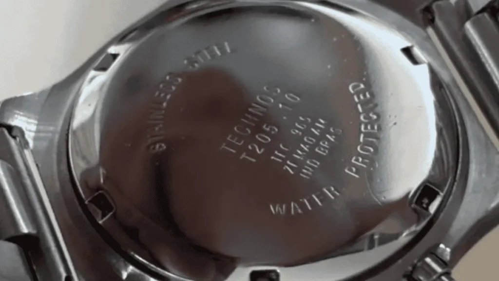 Foto macro da tampa traseira de um relógio Technos original em aço inoxidável, detalhando a referência do modelo e as gravações em baixo relevo.