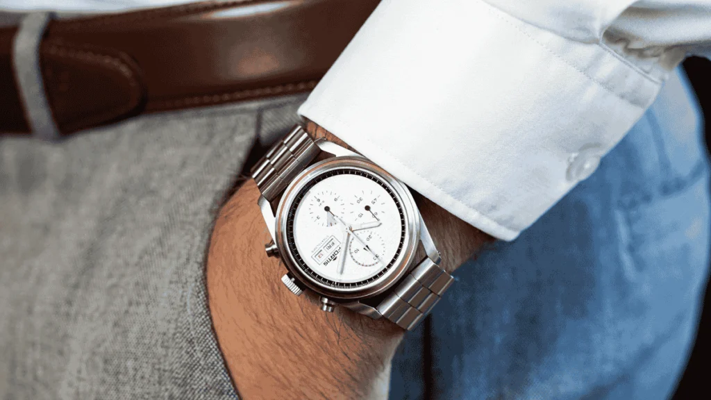 Close de um pulso masculino usando um relógio analógico clássico com pulseira de couro marrom, ajustado sob o punho de uma camisa social branca.